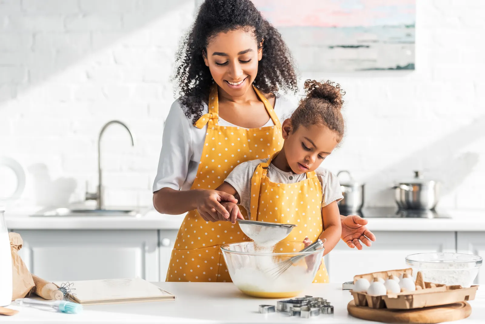 A woman and a child sieving flour into a bowl at a kitchen counter