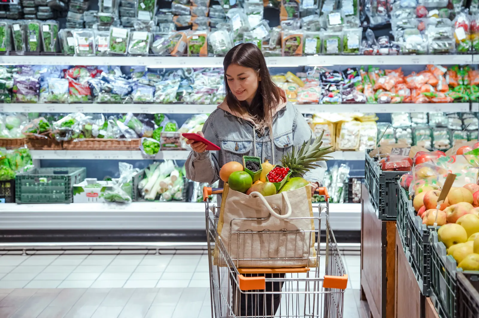 A woman pushing a trolley through a supermarket with her phone in her hand